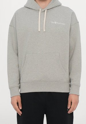 Osoba nosząca jasnoszarą bluzę z kapturem Polo Ralph Lauren z kieszenią z przodu i beżowymi sznurkami, połączoną z czarnymi spodniami.