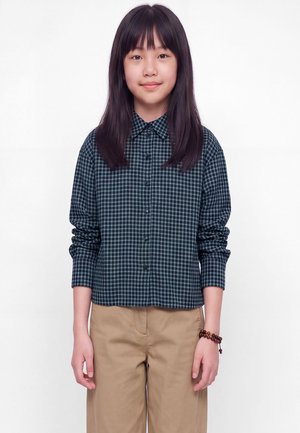 Jeune personne aux longs cheveux foncés portant une chemise à carreaux vert foncé boutonnée et un pantalon beige, debout devant un fond blanc uni.