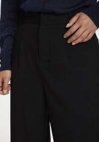 Zwarte broeken met een gladde textuur, voorzien van een geplooide voorkant en een elastische tailleband. Hand rustend op de zijzak.