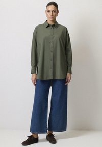 Olivegrön knäppskjorta med lång ärm, kombinerad med vida blå denimbyxor och mörkbruna skor. Slät tygtextur.