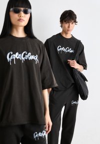 Zwarte oversized t-shirts en broeken met een wit druipend logo. Model houdt een zwarte tas vast. Glad textuur, casual pasvorm. Zonnebril gedragen door het model.