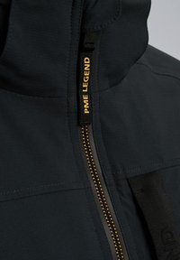 Schwarze Jacke mit hohem Kragen, Reißverschluss mit goldenen Akzenten und einem strukturierten Stoffdesign. Die Markenkennzeichnung ist am Reißverschlussanhänger sichtbar.