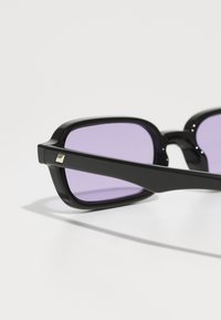 Gafas de sol cuadradas negras con lentes morados, con un diseño elegante y un acento metálico en la pata. Acabado mate con una textura suave.