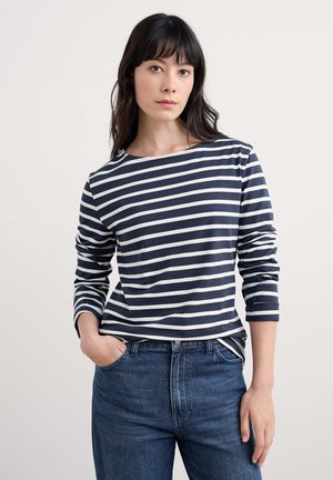 Žena s dlhými tmavými vlasmi, ktorá má na sebe modro-bielu pruhovanú blúzku s dlhými rukávmi a modré džínsy, jedna ruka v vrecku, neutrálne pozadie.