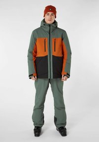 Skijas in groen, oranje en zwart, met een capuchon, twee borstzakken en ritsen. Draagt het met bijpassende broek en zwarte skibotten.