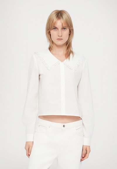 Blouse blanche à manches longues et à coupe courte, dotée d'un large col en dentelle et d'une fermeture par boutons sur le devant. Tissu léger et lisse.