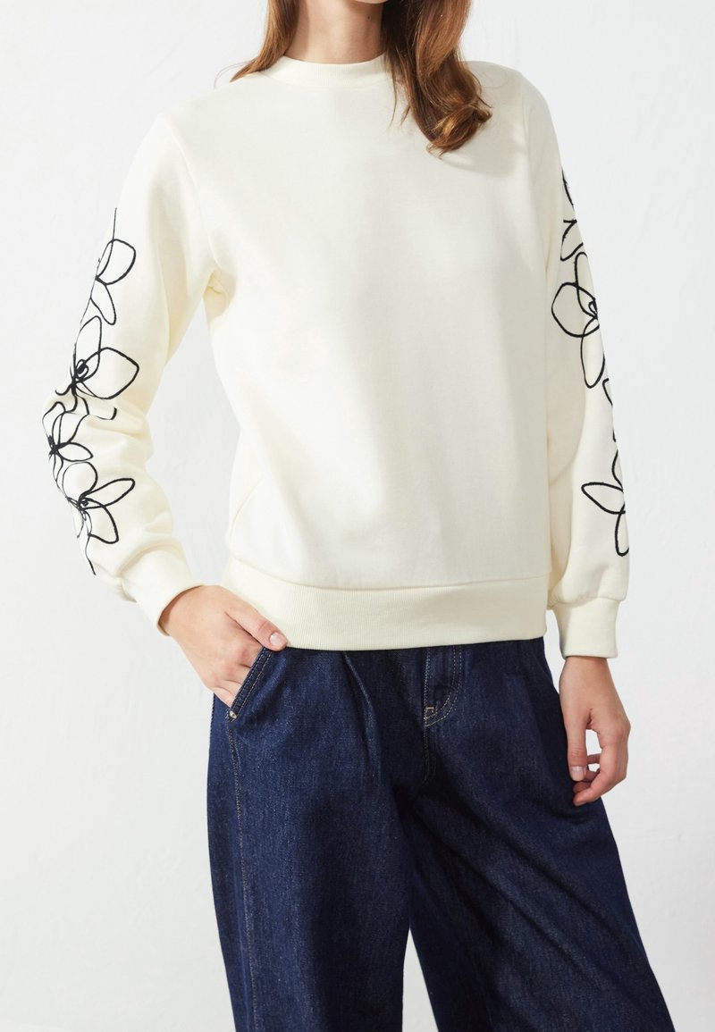 Femme portant un sweat crème avec une broderie florale noire sur les manches et un jean large bleu foncé, debout avec une main dans la poche.