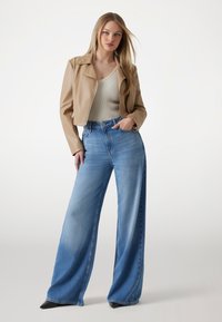 Giacca in pelle beige cropped, top aderente a coste, jeans larghi azzurri, tacchi neri, modella che sta in piedi con sicurezza. Texture morbide, look casual.