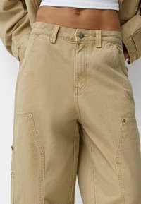 Pantalones cargo caqui de algodón, con una cintura alta, grandes bolsillos laterales y detalle de costura visible. Cierre de botón metálico en la cintura.