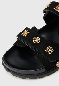 Μαύρο σουέντ σανδάλι με χρυσά διακοσμητικά στοιχεία, διαθέτει δύο ρυθμιζόμενους ιμάντες, στρογγυλή μύτη και υφασμένη μαύρη σόλα.