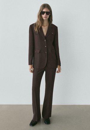 Abito marrone su misura con colletto a risvolto, bottoni doppiopetto e tasche frontali. Indossato con occhiali da sole neri e scarpe nere.