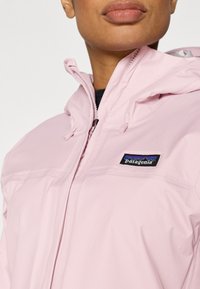 Αδιάβροχο ροζ ανοιχτό μπουφάν με κουκούλα, με φερμουάρ και μπλε λογότυπο Patagonia στο στήθος. Ομαλή υφή και ελαφρύς σχεδιασμός.