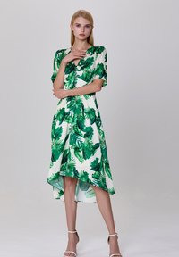 Robe florale présentant des motifs de feuilles vertes sur fond blanc, manches courtes bouffantes et ourlet asymétrique. Texture de tissu lisse.