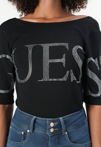 Maglietta nera con design off-shoulder, con grandi lettere in strass argentati che formano la parola "GUESS" sulla parte frontale. Abbinata a jeans in denim blu.