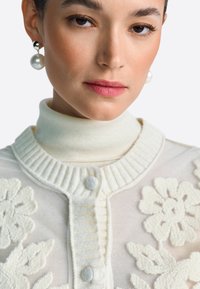 Biely pletený kardigán s kvetinovým vzorom, rebrovaným golierom a zapínaním na gombíky. Má vysoký výstrih a textúrované detaily.