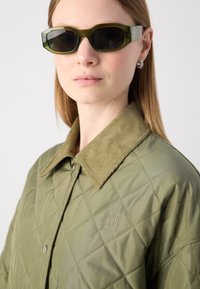 Jachetă matlasată de culoare verde măslinie cu guler din catifea, având un logo pe piept. Purtașul are ochelari de soare întunecați, cu forme unghiulare.