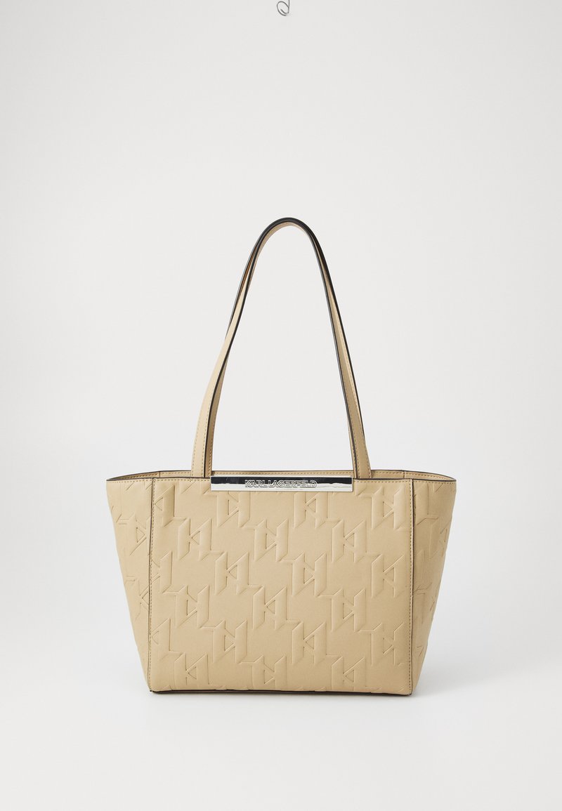 Beige totebag laget av teksturert materiale, med et preget logo-design, korte håndtak og en sølvfarget aksent.