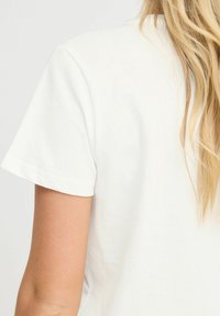 Camiseta de algodón blanca de manga corta, con un cuello redondeado simple y una textura suave. Detalle visible de la costura del hombro.