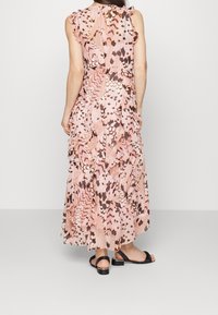 Maxime gown with fließenden Ärmeln, mit einem rosa und braunen Federdesign, aus fließendem Stoff. Trägt schwarze Sandalen mit Riemchen.