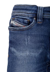 Niebieskie jeansy z denimu z efektem przetarcia, wyposażone w małą przednią kieszeń, ozdobne przeszycia oraz widoczną markową metkę w tylnej kieszeni.