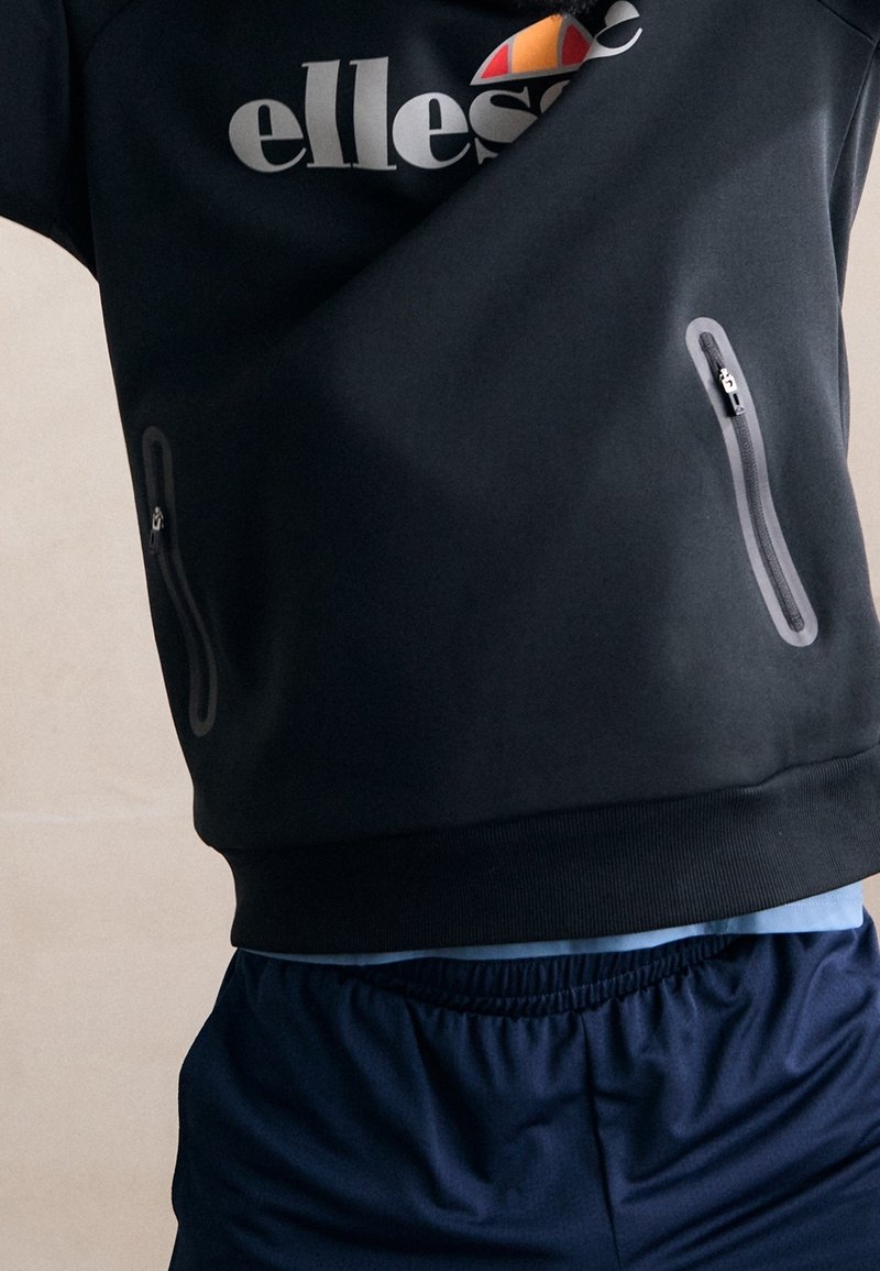 Persona che indossa un felpa nera ellesse con tasche con zip e pantaloni sportivi blu navy, su uno sfondo neutro.