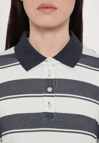 Sötétkék gallérral ellátott pólóing, vízszintes szürke és fehér csíkokkal, világos gombokkal készült gombolással és látható varrási részletekkel.