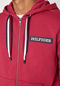 Hoodie zippé rouge en tissu texturé, avec des cordons de serrage contrastants bleu marine et blancs, et un patch noir affichant "HILFIGER" en lettres blanches.
