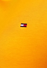 Żółta bawełniana koszulka z krótkimi rękawami z małym, haftowanym logo w czerwonym, białym i niebieskim kolorze, umiejscowionym po lewej stronie w pobliżu klatki piersiowej.