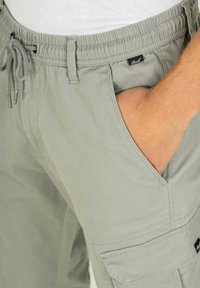 Hellgraue Cargo-Hose mit elastischem Bund, Kordelzug und Seitentaschen. Verfügt über ein gewebtes Etikett an der Vordertasche. Weiche Textur.