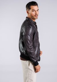 Bruine leren bomberjack met ribgebreide mouwen en zoom, met een soepele textuur en subtiele glans, gedragen over een donker shirt en lichte broek.