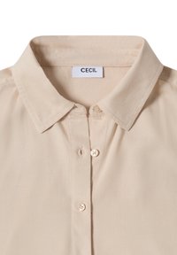 Chemise beige à boutons avec un col classique, en tissu léger, comportant une petite étiquette de marque et deux boutons à l'avant. Texture lisse.