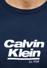 Κοντινό πλάνο μπλε ναυτικού πουκάμισου με λευκό λογότυπο "Calvin Klein" και κείμενο "Est. 1968" τυπωμένο στην περιοχή του στήθους.