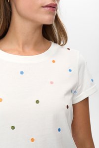 Frau in einem weißen kurzärmeligen Shirt mit mehrfarbigen gestickten Punkten, von Hals bis zur Mitte des Oberkörpers vor einem weißen Hintergrund gezeigt.