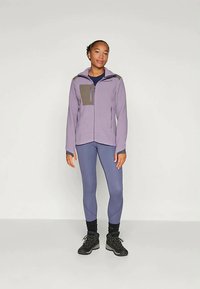 Light purple zip-up jacket with a dark gray chest pocket, paired with fitted blue leggings and black hiking shoes. Smooth texture.