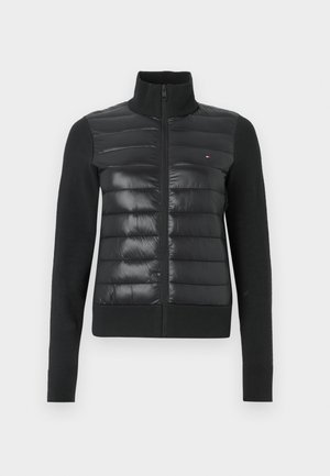 Μαύρο μπουφάν με φερμουάρ μπροστά, που συνδυάζει ένα επενδυμένο, γυαλιστερό πάνω μέρος με πλεκτό κάτω μέρος. Διακριτική λεπτομέρεια λογότυπου στο αριστερό στήθος.