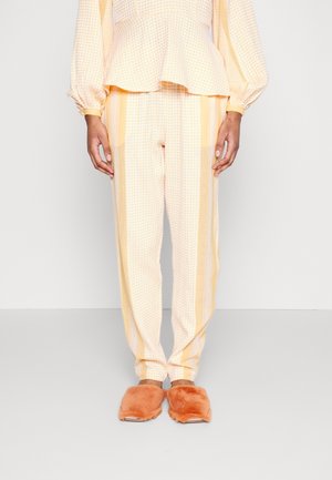 Kobieta ubrana w bluzkę z długim rękawem w bladoróżową kratę oraz dopasowane spodnie z pionowymi paskami, do tego pomarańczowe, puszyste kapcie.