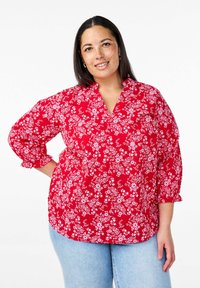 Blusa rossa floreale con motivi bianchi, maniche a tre quarti, scollatura a V e polsini arricciati; abbinata a jeans azzurri chiari.