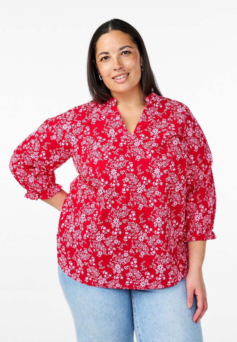 Blusa rossa floreale con motivi bianchi, maniche a tre quarti, scollatura a V e polsini arricciati; abbinata a jeans azzurri chiari.