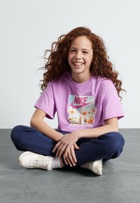 Camiseta morada de Nike con gráfico floral y lema, combinada con pantalones de chándal azul marino. Zapatillas blancas con acentos crema. Pose sentada sobre una superficie gris.