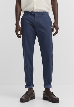 Uomo che indossa pantaloni blu scuro risvoltati, scarpe in pelle marrone scuro e camicia chiara con bottoni e trama.