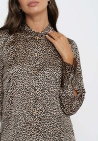 Blusa con stampa leopardata realizzata in tessuto morbido, dotata di colletto, chiusura frontale con bottoni e maniche arrotolate con dettagli a tab. Toni di beige e nero.
