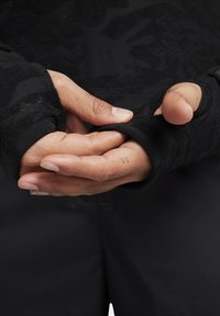 Černé bezprsté rukavice s texturovanou tkaninou a elastickým manžetou. Ruce jsou sepnuté dohromady, přičemž je vidět tetování na zápěstí.