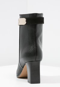 Bota de cuero negro para tobillo con tacón medio y bloque. Presenta un detalle de hebilla plateada y textura suave, diseñada para una silueta ajustada.