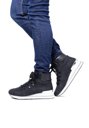 Zapatillas altas de ante negras con suelas blancas, con ojales y cordones de metal. Llevadas con jeans ajustados azules para un look casual.