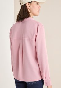 Helles pinkes Langarmbluse mit einem Rückenfalte, geknöpften Manschetten und subtilen Stickdetails, kombiniert mit dunklen Jeans.