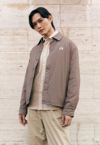 Brown lightweight jacket with a collared neck, snap buttons, and a patch logo, layered over a cream button-up shirt and light trousers.
