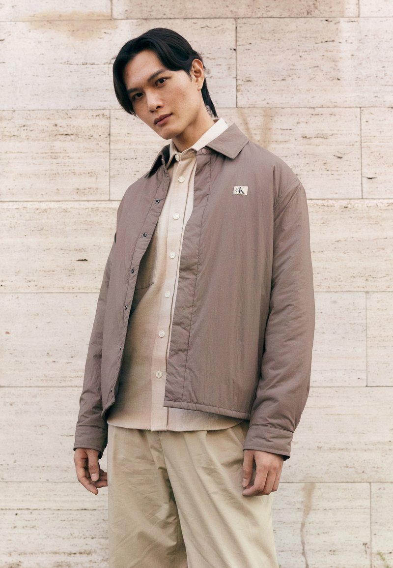 Brown lightweight jacket with a collared neck, snap buttons, and a patch logo, layered over a cream button-up shirt and light trousers.