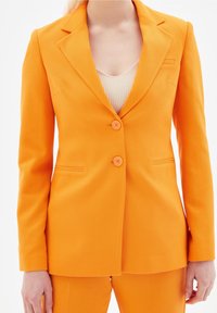 Pomarańczowa dopasowana marynarka z wyciętymi klapami, dwoma guzikami z przodu i bocznymi kieszeniami. Teksturowana tkanina o gładkim wykończeniu. Podszewka wewnętrzna.