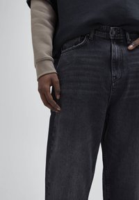 Vaqueros de denim gris oscuro con un corte relajado, que cuentan con un cierre de botón y un ligero desgastado. Acompañados de una camisa de manga larga beige.
