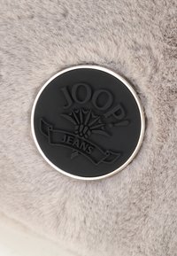 Grå faux fur materiale med et cirkulært sort plastlogo med "JOOP! JEANS" i præget skrift og sølvkant.